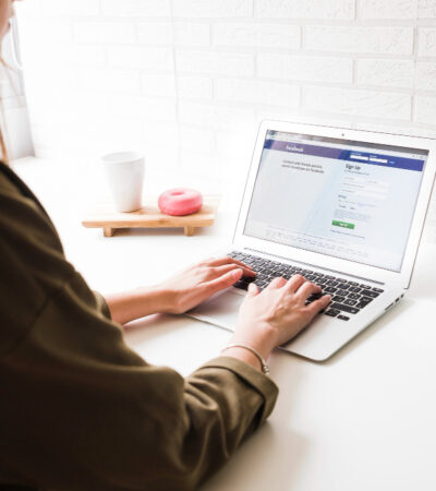 A person is working on a laptop showing a social media sign-up page, with a cup, a doughnut on a small tray, and a potted cactus on the table, creating a casual work environment.